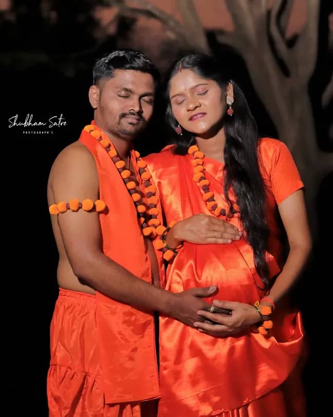 Couple dressed in matching orange traditional attire standing close together, with the woman holding her pregnant belly and the man gently placing his hand over hers. They are adorned with orange garlands and jewelry, standing outdoors at night with a tree in the background. The mood is warm and celebratory. Text in the image reads Shubham Satre Photographs.